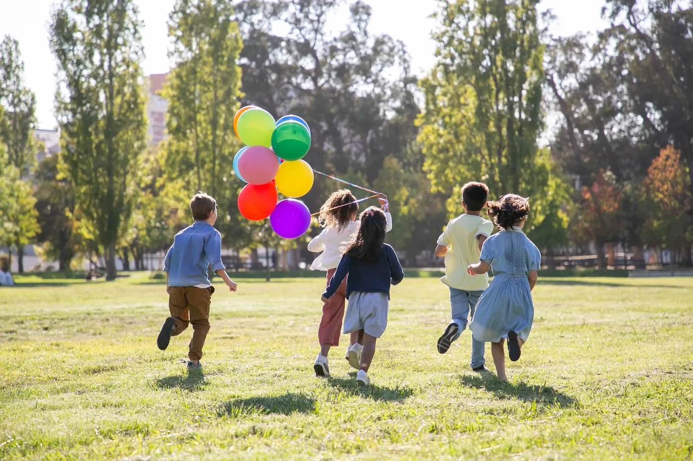 Copii aleargă cu baloane colorate printr-un parc, fotografiați din spate, într-o zi însorită